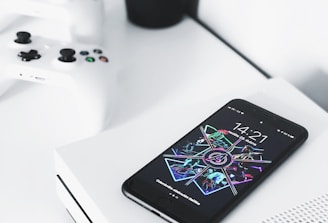 A smartphone with a colorful and futuristic display lies on top of a white gaming console. Two white gaming controllers are placed nearby on a white surface. In the background, there is a small black pot containing green, leafy plants, adding contrast and a touch of nature to the modern setup.