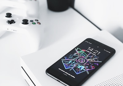 A smartphone with a colorful and futuristic display lies on top of a white gaming console. Two white gaming controllers are placed nearby on a white surface. In the background, there is a small black pot containing green, leafy plants, adding contrast and a touch of nature to the modern setup.