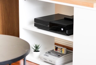 A black gaming console is situated on the middle shelf of a modern, white wooden entertainment unit. Below it, an older version of a gaming console sits with a game cartridge inserted. A small potted plant adds a touch of greenery next to the vintage console. The top of the unit is made of a light wood, and a flat-screen monitor is partly visible on it. The setup is minimalistic and clean, with a round black table in the foreground.