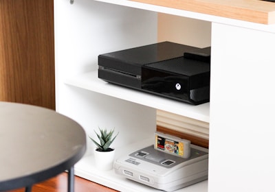 A black gaming console is situated on the middle shelf of a modern, white wooden entertainment unit. Below it, an older version of a gaming console sits with a game cartridge inserted. A small potted plant adds a touch of greenery next to the vintage console. The top of the unit is made of a light wood, and a flat-screen monitor is partly visible on it. The setup is minimalistic and clean, with a round black table in the foreground.