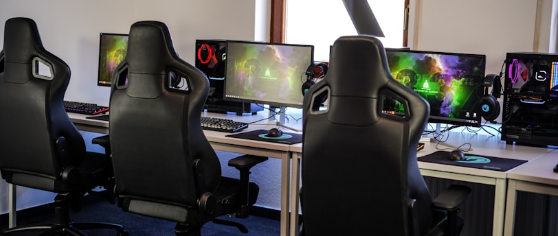 A row of gaming stations equipped with large monitors, gaming chairs, keyboards, and headphones. The setup is organized and ready for gaming, featuring a colorful screen display. The room has a modern and tech-savvy ambiance.