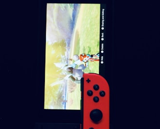 A gaming console with a screen displaying a video game scene, accompanied by two controllers, one colored red and the other blue. The devices are set against a dark backdrop, emphasizing the vibrant colors of the controllers.