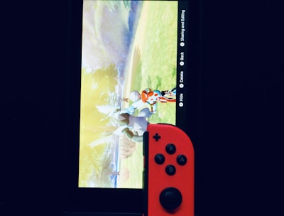 A gaming console with a screen displaying a video game scene, accompanied by two controllers, one colored red and the other blue. The devices are set against a dark backdrop, emphasizing the vibrant colors of the controllers.
