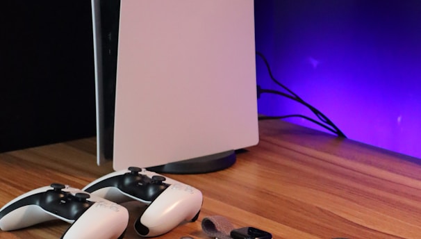 A sleek, modern setup featuring a gaming console with two controllers on a wooden surface. Nearby are a smartphone, smartwatch, tablet with a vivid screen, wireless earbuds, and a stylus. The background is illuminated with a soft purple glow.