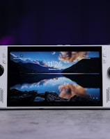 A handheld gaming console with a central screen displaying a scenic wallpaper of mountains and a lake, flanked by controller buttons and joysticks on both sides. The lighting around is dim, with purplish hues in the background, creating a modern and elegant atmosphere.