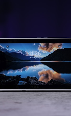 A handheld gaming console with a central screen displaying a scenic wallpaper of mountains and a lake, flanked by controller buttons and joysticks on both sides. The lighting around is dim, with purplish hues in the background, creating a modern and elegant atmosphere.