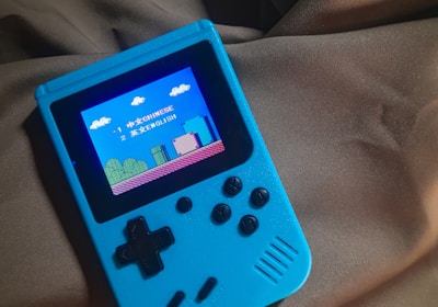 A handheld gaming device with a bright blue casing rests on a soft, dark fabric background. The screen displays a pixelated video game scene with options to select either Chinese or English language. The layout includes a classic D-pad and buttons, reminiscent of retro gaming consoles.